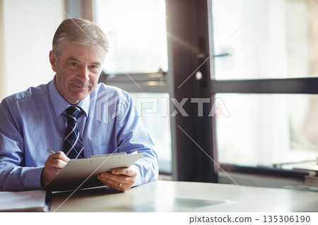 Middle-aged man sitting at desk in modern office holding clipboard and pen taking notes, copy space Middle-aged man sitting at desk in modern office holding clipboard and pen taking notes, copy space 135306190