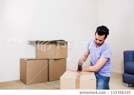 Male kneeling sealing box with packing tape next to blue couch at room corner, copy space Male kneeling sealing box with packing tape next to blue couch at room corner, copy space 135306195