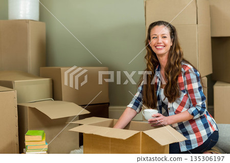 Woman kneeling at home packing cardboard boxes while holding ceramic mug next to books, copy space Woman kneeling at home packing cardboard boxes while holding ceramic mug next to books, copy space 135306197