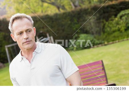 Middle-aged man wearing white polo shirt standing in backyard garden by trimmed hedge and red bench 135306199