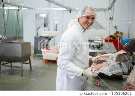 Mid adult man wearing hairnet handling meat cuts on metal table at meat plant, copy space 135306201
