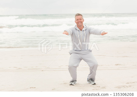 Mature man squatting with arms extended wearing light gray hoodie and sweatpants on sandy shoreline 135306208