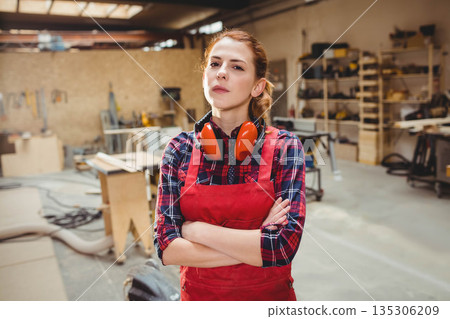 Female woodworker wearing red apron and earmuffs, standing amid workbenches and lumber in workshop 135306209