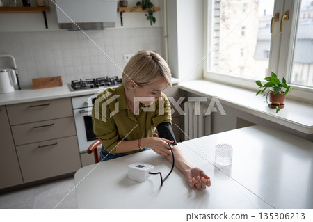 Middle aged woman sits at table measuring blood pressure with tonometer, seasonal health problems. 135306213