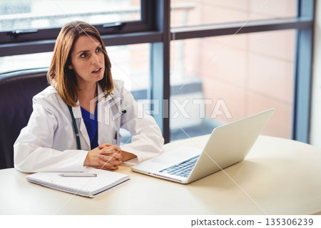 Woman doctor wearing scrubs and stethoscope sitting at desk reviewing notes on laptop and notebook 135306239