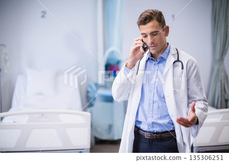 Male doctor in his 30s talking on smartphone while gesturing between hospital ward beds, copy space 135306251