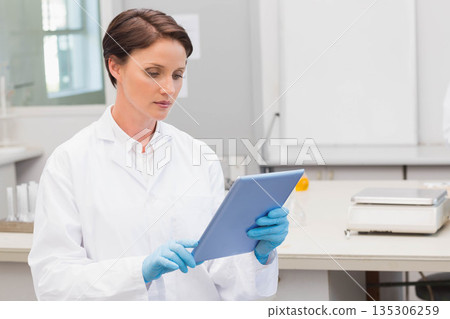 Gloved woman scientist in lab coat reviewing data on tablet at bench with balance, copy space Gloved woman scientist in lab coat reviewing data on tablet at bench with balance, copy space 135306259