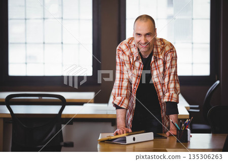 Man leaning forward on desk and smiling at camera in modern office space with tablet, binder Man leaning forward on desk and smiling at camera in modern office space with tablet, binder 135306263
