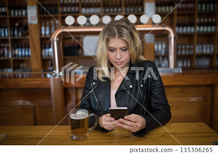 Woman sitting at wooden table in pub using smartphone and tapping screen near mug of beer 135306265