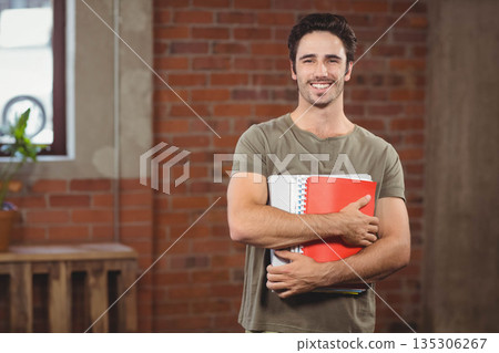 Male student smiling while holding spiral notebooks and red folder in brick-walled room with plant Male student smiling while holding spiral notebooks and red folder in brick-walled room with plant 135306267