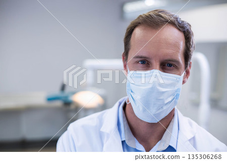 Male dentist wearing surgical mask and lab coat checking controls with dental lamp in clinic Male dentist wearing surgical mask and lab coat checking controls with dental lamp in clinic 135306268