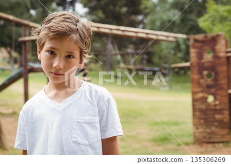 School-age boy standing on grass near wooden climbing frame and metal bars at playground School-age boy standing on grass near wooden climbing frame and metal bars at playground 135306269