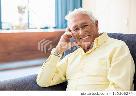 Senior man sitting on sofa in living room, wearing eyeglasses and wristwatch beside wooden console 135306274
