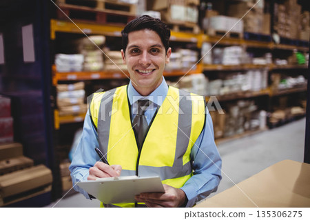 Man wearing high-visibility vest in warehouse shelving holding clipboard, pen inspecting inventory Man wearing high-visibility vest in warehouse shelving holding clipboard, pen inspecting inventory 135306275