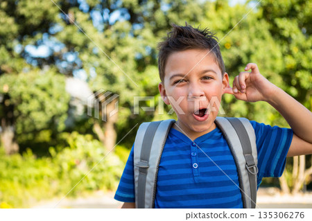 Boy pulling ear and making playful face while standing outdoors in park with backpack, copy space Boy pulling ear and making playful face while standing outdoors in park with backpack, copy space 135306276