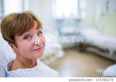 Senior woman reclining on hospital bed near empty bed in patient room wearing patterned gown Senior woman reclining on hospital bed near empty bed in patient room wearing patterned gown 135306278