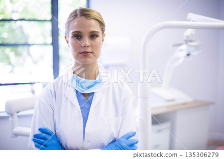 Adult female dentist standing with crossed arms in dental clinic near window and overhead lamp 135306279