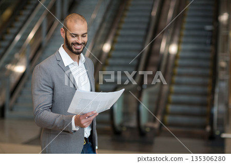 Man standing in large lobby reading folded newspaper near parallel escalators with polished floor 135306280