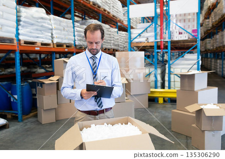 Male supervisor in office attire in warehouse holding clipboard inspecting box with packing peanuts Male supervisor in office attire in warehouse holding clipboard inspecting box with packing peanuts 135306290