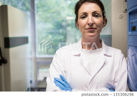 Mid adult female scientist wearing lab coat and blue gloves standing beside freezer in laboratory 135306296