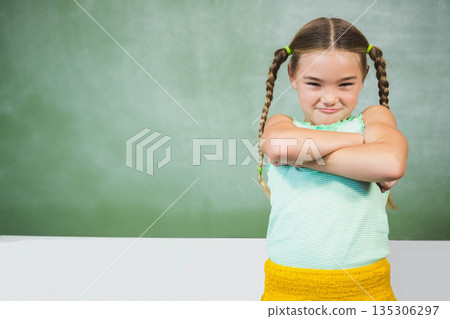 Female child standing with crossed arms in classroom at green chalkboard by white desk, copy space 135306297