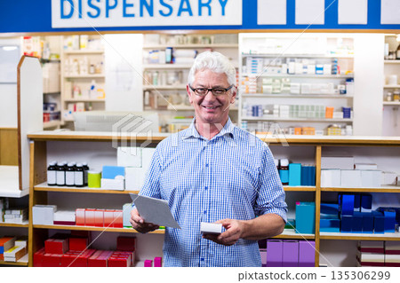 Middle-aged pharmacist checking prescription pad in dispensary behind counter by medication shelves 135306299