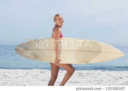 Mid adult woman wearing red swimsuit walking along white sandy beach holding longboard surfboard Mid adult woman wearing red swimsuit walking along white sandy beach holding longboard surfboard 135306307