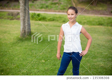 female child standing on grassy lawn in park beside mature tree trunk and dirt path 135306308