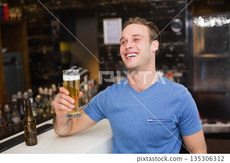 Man leaning at bar counter and holding beer glass with foam beside brown bottle and shelves Man leaning at bar counter and holding beer glass with foam beside brown bottle and shelves 135306312