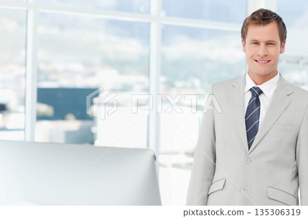 Man smiling while standing in suit next to office desk computer monitor in workspace, copy space 135306319