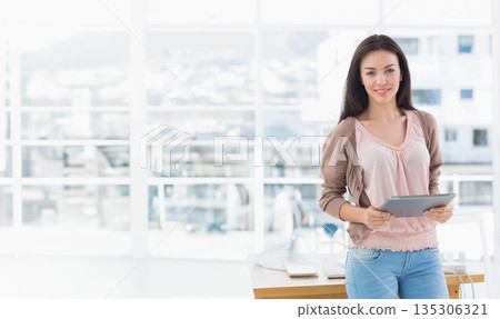 Businesswoman holding tablet in office by windows with desk, keyboard, laptop and mouse, copy space Businesswoman holding tablet in office by windows with desk, keyboard, laptop and mouse, copy space 135306321