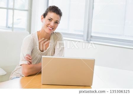 Woman sitting on cushioned chair at wooden office desk using laptop under large windows, copy space 135306325