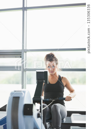 Woman pulling rower handle while monitoring digital screen in gym by windows in sportswear 135306336