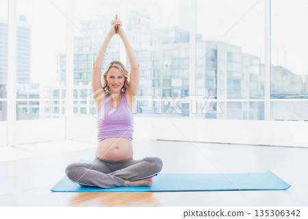 Pregnant woman stretching arms overhead in prenatal yoga pose on blue mat in studio with windows Pregnant woman stretching arms overhead in prenatal yoga pose on blue mat in studio with windows 135306342