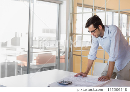Adult male professional leaning over table studying blueprints and using calculator in meeting room Adult male professional leaning over table studying blueprints and using calculator in meeting room 135306348