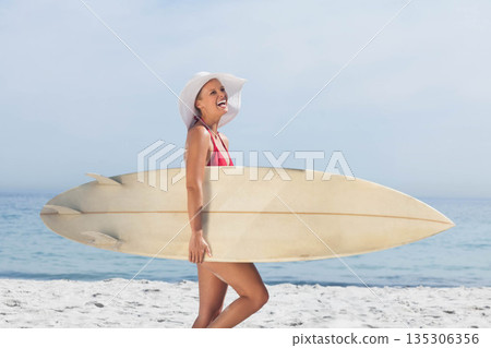 Woman walking along shoreline in red swimsuit and white sun hat holding long surfboard Woman walking along shoreline in red swimsuit and white sun hat holding long surfboard 135306356