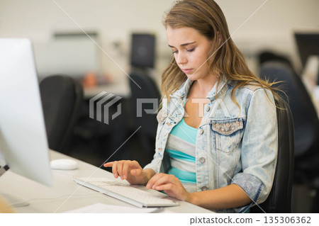 Woman wearing denim jacket typing on computer keyboard and using mouse at shared office workspace 135306362