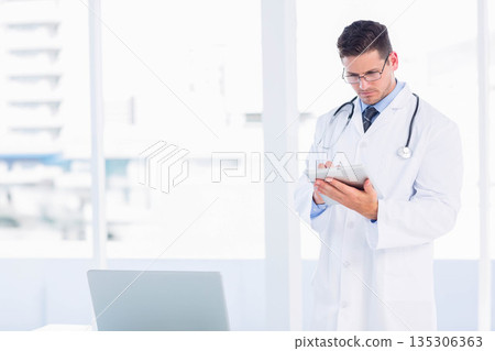 Male physician holding tablet reviewing patient data at office desk with laptop, copy space Male physician holding tablet reviewing patient data at office desk with laptop, copy space 135306363