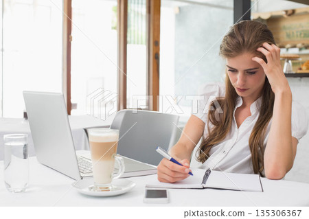 Woman writing in spiral notebook with pen at coffee shop table beside laptop, copy space Woman writing in spiral notebook with pen at coffee shop table beside laptop, copy space 135306367