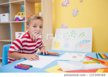 Boy wearing striped t-shirt drawing at blue table in art corner with pencils, crayons, copy space Boy wearing striped t-shirt drawing at blue table in art corner with pencils, crayons, copy space 135306368