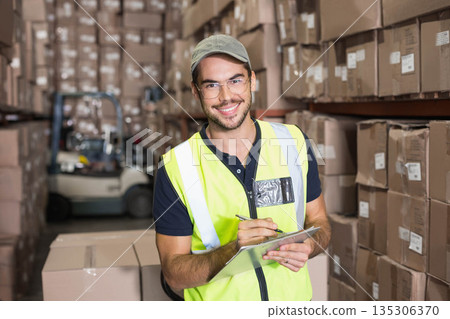 Man wearing safety vest standing in warehouse holding clipboard and pen near forklift and boxes Man wearing safety vest standing in warehouse holding clipboard and pen near forklift and boxes 135306370