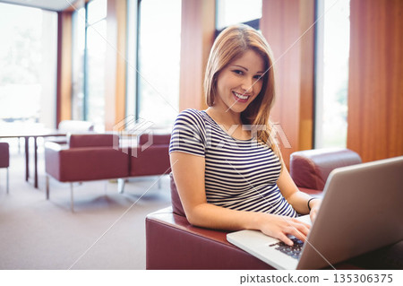 Female student sitting in leather armchair in study lounge near large windows working on laptop Female student sitting in leather armchair in study lounge near large windows working on laptop 135306375