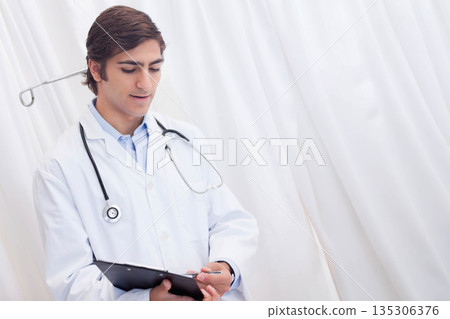 Male doctor in lab coat writing patient notes in clinic with clipboard and stethoscope, copy space Male doctor in lab coat writing patient notes in clinic with clipboard and stethoscope, copy space 135306376