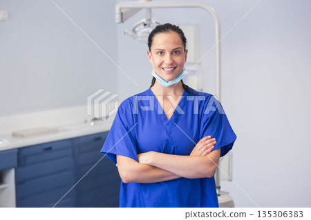 Female dentist standing in dental clinic wearing blue scrubs with mask under chin and overhead lamp Female dentist standing in dental clinic wearing blue scrubs with mask under chin and overhead lamp 135306383