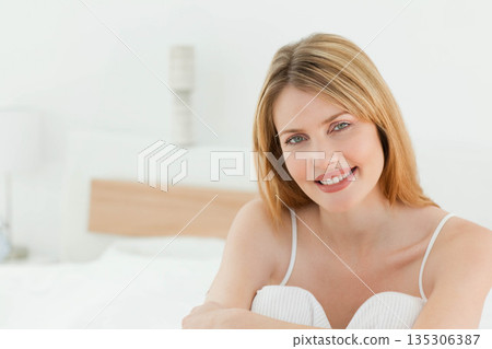 Woman leaning forward on bed in bright bedroom with wooden headboard and wall lamp, copy space Woman leaning forward on bed in bright bedroom with wooden headboard and wall lamp, copy space 135306387