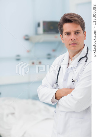 Male doctor standing with crossed arms wearing lab coat and stethoscope in patient room, copy space 135306388