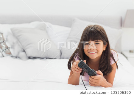 Girl lying on bed holding black wired game controller among gray and white pillows in bedroom Girl lying on bed holding black wired game controller among gray and white pillows in bedroom 135306390