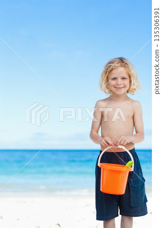 Boy standing on beach wearing swim shorts and holding orange bucket with green shovel, copy space Boy standing on beach wearing swim shorts and holding orange bucket with green shovel, copy space 135306391
