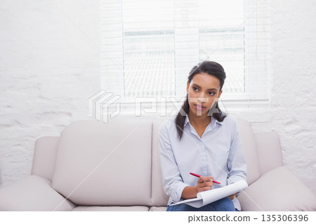 African American woman holding red pen writing in notepad while sitting on sofa in living room 135306396