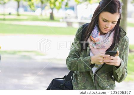 Woman standing on walkway in park in camo jacket, holding smartphone and black bag, copy space 135306397